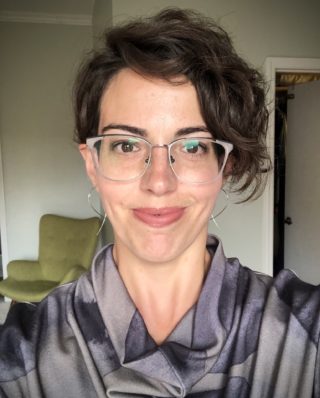 Headshot of a woman with short wavy hair and fair skin, wearing glasses and earrings, with a soft smile and warm eyes.