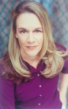 Carolyn, a caucasian woman, is shown in 3/4 leaning into the camera wearing a  magenta blouse, with a diffuse urban background behind her.