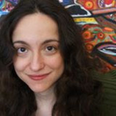 Nadia, a Jewish woman with dark hair, stands in front of a multi-colored painting.